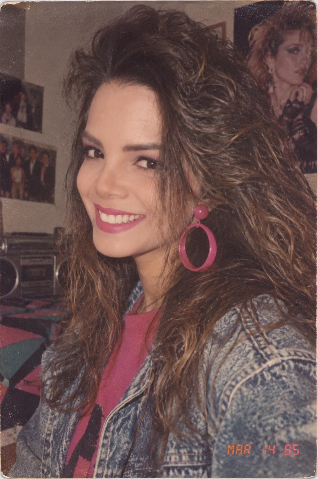 A playful retro-style snapshot: smiling woman with big 80s hair, pink hoop earrings and an acid-wash denim jacket